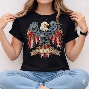 May include: Black t-shirt with a detailed eagle graphic in patriotic colours. The eagle's wings are decorated with stars and stripes, and the text "EST. 1776" is on a banner. The shirt is made of a soft material. The design is reminiscent of the American flag.