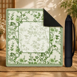 May include: A square mat featuring a cream background with a green floral pattern and a geometric border. The mat has a solid black underside and is shown with a matching black cylindrical carrying case with a shoulder strap for transport.
