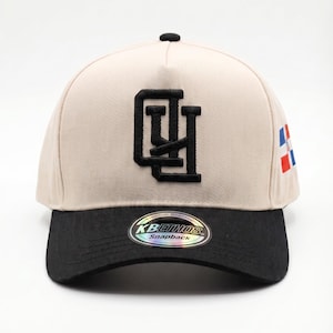 May include: A beige and black snapback hat with a black embroidered logo and a small flag design on the side. The hat features a curved brim and a snap closure at the back.