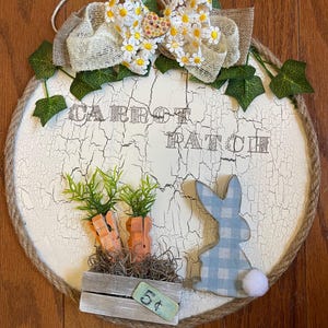 May include: Round, distressed white wooden sign with the words "CARROT PATCH" in brown lettering. The sign features a burlap bow with daisy accents, faux carrots, and a blue gingham bunny with a white pom-pom tail. A small sign reads "5¢".