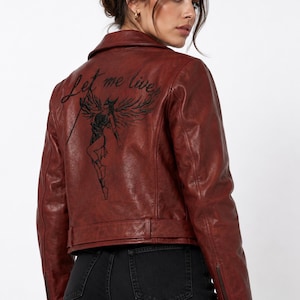 May include: A dark red leather jacket featuring a black graphic print on the back of a winged figure holding a staff, accompanied by the text Let me live in a cursive script. The jacket has a collared neckline, long sleeves with zips, and a belt.