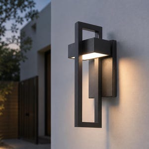 May include: A contemporary black outdoor wall light. The rectangular light features a geometric design with a boxy top and a vertical rectangular frame. It's mounted on a light grey wall, emitting a warm glow. Ideal for exterior lighting.