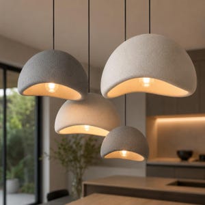 Könnte beinhalten: Vier Pendelleuchten mit einer abgerundeten, kuppelförmigen Form. Die Lampenschirme sind in Grau- und Off-White-Tönen gehalten und haben eine strukturierte, filzartige Optik. Jede Leuchte hat eine warme, leuchtende Glühbirne, die von unten sichtbar ist. Die Leuchten hängen an schwarzen Kabeln.