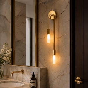 May include: A gold-toned wall sconce with two vertical light fixtures. The sconce has a circular base and a curved arm that holds the cylindrical lights. The lights emit a warm glow, and the fixture is mounted on a marble-patterned wall.