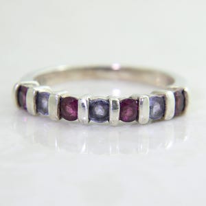 May include: A silver band ring featuring a row of alternating round-cut purple and deep red gemstones. The stones are set in a bar-style setting, with each individual gem separated by a small vertical silver bar, creating a clean and structured look.