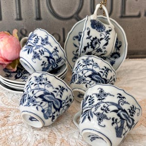 May include: A set of white porcelain teacups and saucers featuring a blue onion pattern with intricate floral and vine motifs. The cups have scalloped edges and curved handles, with some pieces stacked and others arranged to display the blue designs.