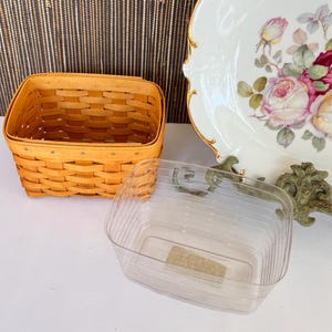 May include: A small, square, woven basket in a light brown color sits next to a clear plastic container. A decorative plate with a floral design and gold trim is also visible.