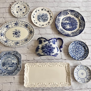 May include: A collection of ceramic tableware including a blue and white floral oval platter, a pitcher, several round plates with landscape and floral patterns, a square plate with a building illustration, and a rectangular tray with lace-like edges.