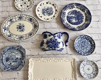 May include: A collection of ceramic tableware including a blue and white floral oval platter, a pitcher, several round plates with landscape and floral patterns, a square plate with a building illustration, and a rectangular tray with lace-like edges.