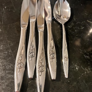 May include: A set of vintage silverware, including four butter knives and one spoon. The silverware is silver-colored with a floral rose design on the handles. The spoon and knives are arranged on a dark green marble surface.