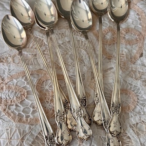 May include: A set of seven silver-toned metal spoons with long, slender handles. Each handle features an ornate, embossed floral design at the base and a small engraved letter. The spoons have polished, reflective oval bowls and smooth, tapered stems.
