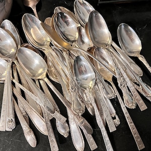 May include: A collection of antique silver spoons, forks, and serving utensils. The silverware has ornate floral and geometric patterns. The spoons have oval bowls and long handles. The items are arranged on a dark surface.