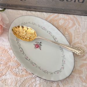 May include: An oval white porcelain dish featuring a central pink rose and a delicate green and pink floral border, paired with a gold-toned metal spoon that has an embossed fruit design on the bowl and a twisted handle pattern.
