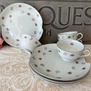 May include: Set of four white teacups and saucers with a delicate floral pattern. The cups and saucers have a scalloped edge and a gold trim. The pattern features small pink roses and gray leaves.