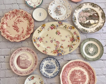 May include: A collection of fifteen ceramic plates and saucers of varying sizes and designs, including floral patterns, landscape scenes, and solid colors. The assortment features scalloped and smooth edges, with colors ranging from red, blue, and green to pink and cream.