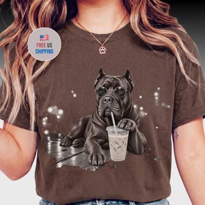 May include: A brown short-sleeved t-shirt featuring a graphic of a black dog wearing round glasses and holding a clear plastic cup with a straw. The dog is positioned against a dark, textured background with light bokeh effects.