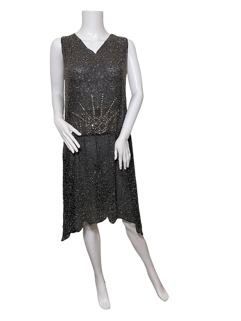 May include: A sleeveless black beaded dress with a sunburst design in the center of the bodice. The dress has a V-neck and a high waist.