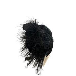 May include: A black, decorative hat with a cluster of long, black feathers and a sequined design. The hat has a rounded shape and is likely made of fabric. The feathers cascade down the side, adding a dramatic flair to the headwear.