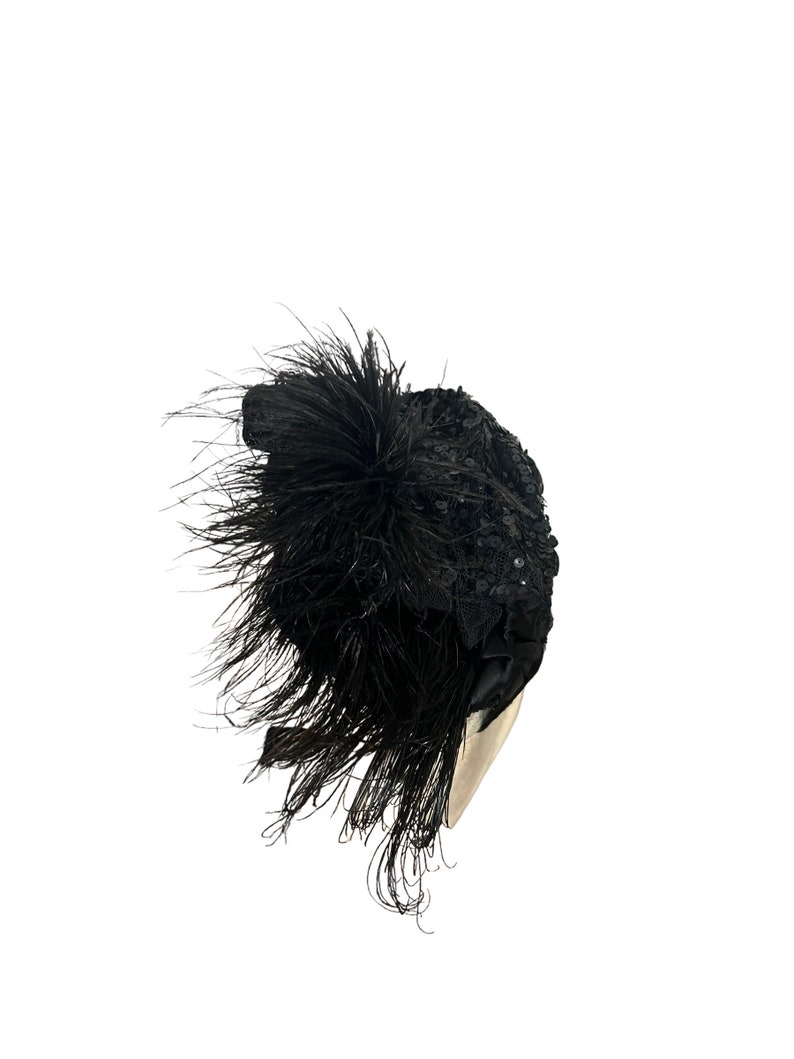 May include: A black, decorative hat with a cluster of long, black feathers and a sequined design. The hat has a rounded shape and is likely made of fabric. The feathers cascade down the side, adding a dramatic flair to the headwear.