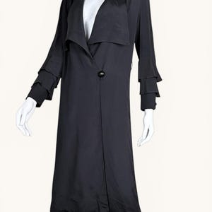 Peut inclure: Un long manteau noir avec un col drapé et une fermeture à bouton unique. Les manches présentent des poignets à volants superposés. Le manteau est présenté sur un mannequin sur fond blanc, mettant en valeur son design élégant.