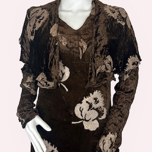 May include: A brown velvet dress and bolero jacket set. The dress has a V-neck and long sleeves, with embroidered floral patterns in a lighter colour. The bolero has a ruffled edge and matching floral embroidery.
