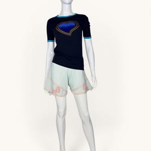 May include: A dark navy short-sleeved knit top with a bright blue heart graphic on the chest and light blue trim at the neck and sleeves, paired with pale mint green shorts featuring a scalloped hem and delicate pink floral lace detailing.