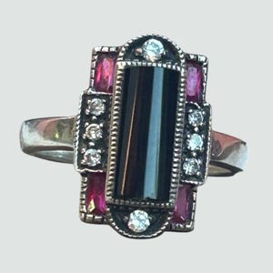 Puede incluir: Anillo plateado con una piedra rectangular de ónix negro, acentuado con pequeñas gemas blancas y piedras rosas rectangulares. El anillo tiene un diseño Art Déco.