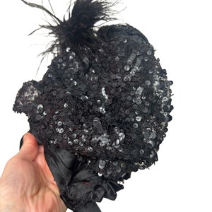 May include: A black sequined hat with a cluster of black feathers. The hat is covered in small, shiny sequins and has a satin ribbon tie. The hat is held by a hand against a white background.