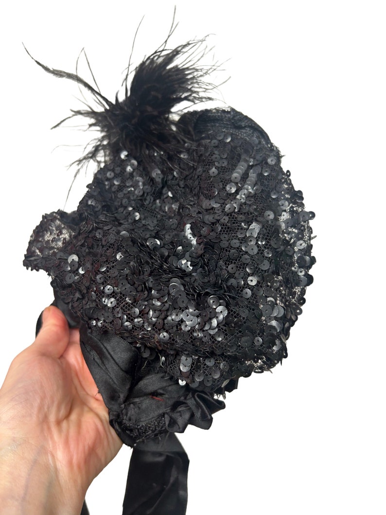 May include: A black sequined hat with a cluster of black feathers. The hat is covered in small, shiny sequins and has a satin ribbon tie. The hat is held by a hand against a white background.