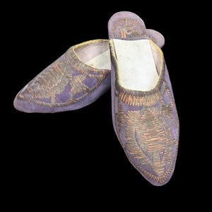 May include: A pair of purple suede slippers with detailed copper and brown embroidery. The slippers have a pointed toe and a soft interior. The design features a leaf-like pattern. The slippers are likely to be around 25 cm in length.