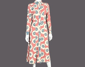 May include: A long-sleeved dress with a floral pattern. The dress features a coral background with white and blue floral designs. The dress has a button-up front and a collared neckline. The dress is displayed on a mannequin.