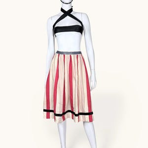 Puede incluir: Conjunto de estilo vintage en un maniquí. El conjunto incluye un top negro, una falda a rayas con rayas verticales rojas y crema y un sombrero negro. La falda tiene una banda negra en el dobladillo. El conjunto se exhibe en un maniquí blanco.