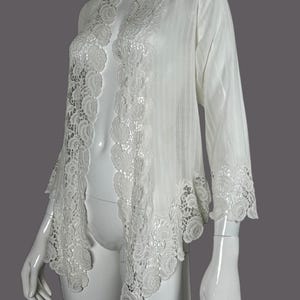 Puede incluir: Chaqueta blanca de frente abierto con ribete de encaje floral. La chaqueta presenta mangas largas y una tela delicada y texturizada. Los detalles de encaje son prominentes a lo largo de los bordes delanteros y los puños de las mangas, creando un look elegante y femenino.