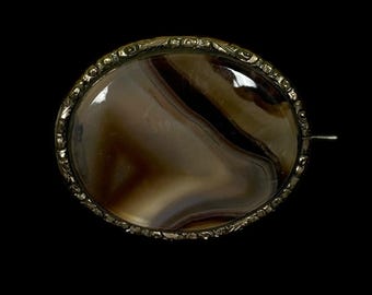 May include: Oval-shaped antique brooch with a polished agate stone. The stone features swirling bands of brown and cream colors. The brooch is set in a decorative, ornate metal frame with a pin closure on the back. The background is black.