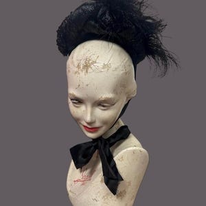 May include: A black hat with feather accents sits atop a mannequin head. The hat has a rounded crown and a wide brim. A black bow is tied around the mannequin's neck. The mannequin has red lipstick.