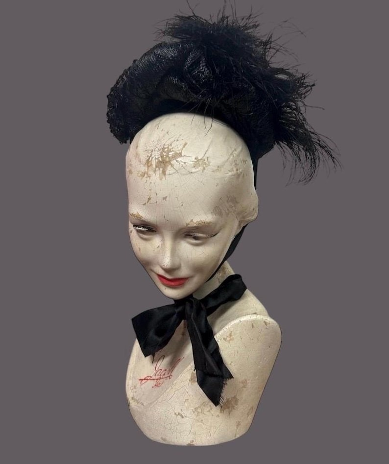 May include: A black hat with feather accents sits atop a mannequin head. The hat has a rounded crown and a wide brim. A black bow is tied around the mannequin's neck. The mannequin has red lipstick.