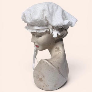 May include: A white bonnet featuring a subtle swiss dot texture and a delicate lace trim along the edge. The head covering includes a smooth fabric band with a small bow detail on the side and long, thin fabric ties hanging down from the brim.