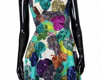 May include: A sleeveless mini dress featuring a white base with a light blue watercolor-style print and large, textured floral-shaped patches covered in multicolored sequins, including shades of purple, green, gold, and blue, with thin straps.