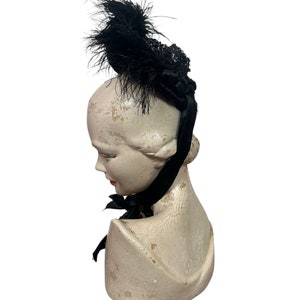 May include: A black vintage hat with a feather and lace design. The hat is adorned with a black ribbon and is displayed on a mannequin head. The mannequin head is painted in a cream color.