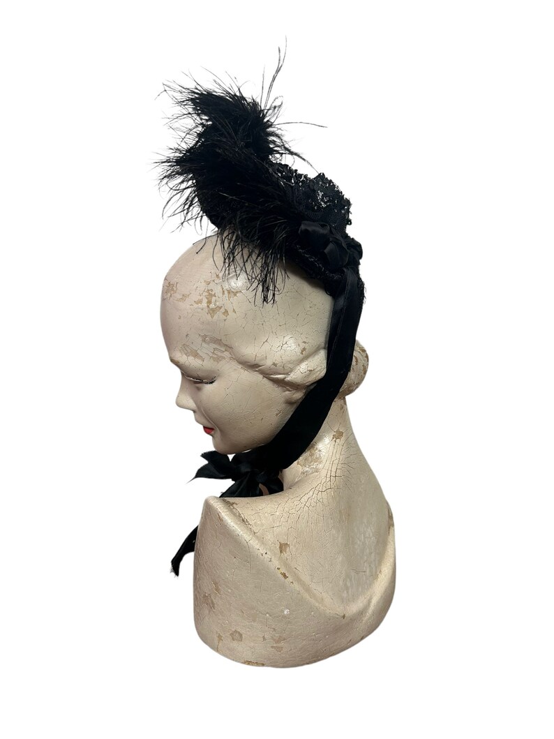 May include: A black vintage hat with a feather and lace design. The hat is adorned with a black ribbon and is displayed on a mannequin head. The mannequin head is painted in a cream color.