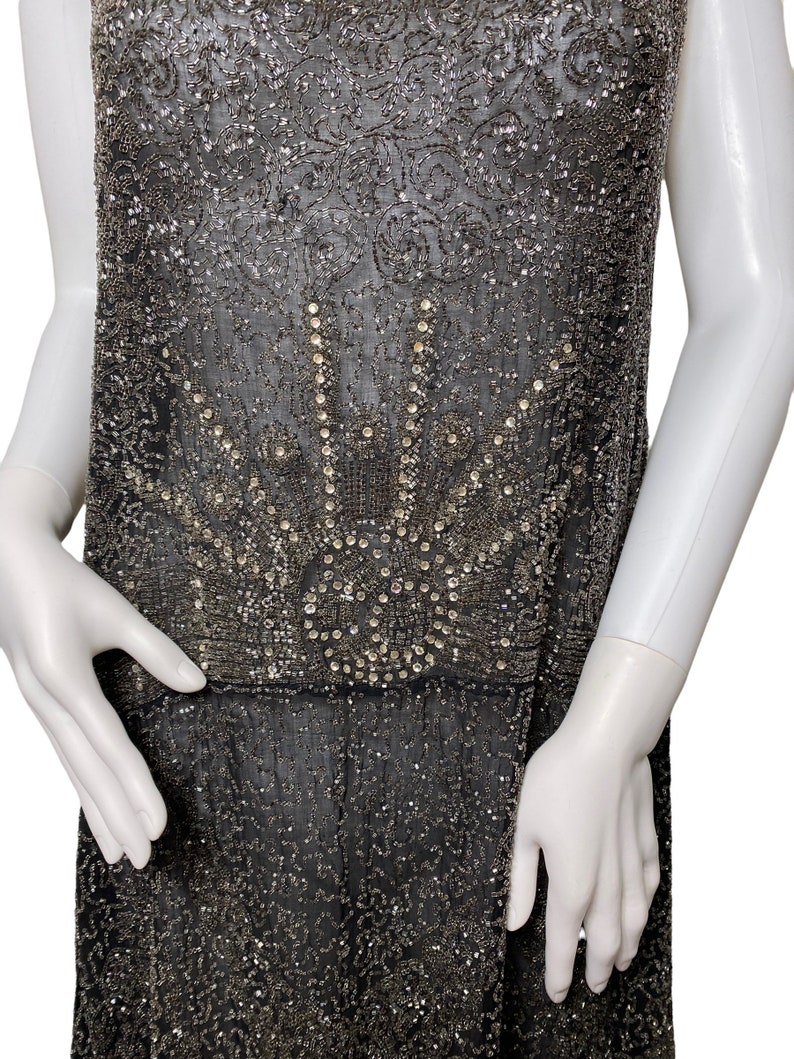 May include: A black beaded dress with a silver and crystal design. The dress has a sleeveless design and a loose fit.