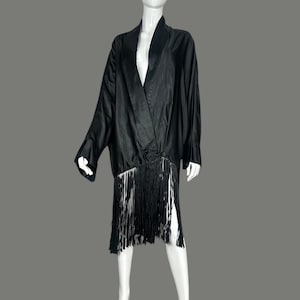 May include: A black satin dress with a shawl collar and long sleeves. The dress features a fringe hemline and a slit on the side. The dress is displayed on a mannequin against a gray background.