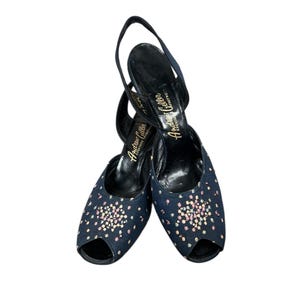 May include: A pair of navy blue slingback heels with an open toe design. The heels are embellished with small, colorful, iridescent sequins. The brand name "Andrew Geller" is printed in gold on the insole.