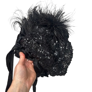 May include: Black sequined hat with black feathers and satin ribbons. The hat is held by a hand against a white background. The hat is covered in small, shiny sequins.