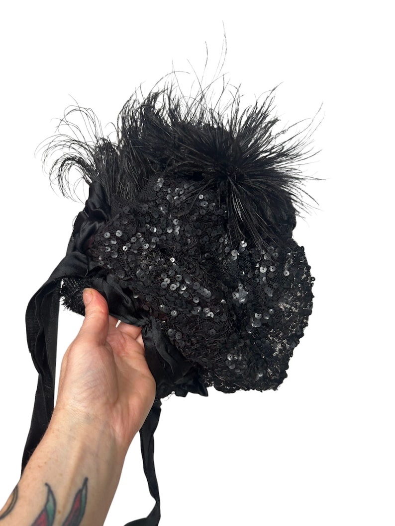 May include: Black sequined hat with black feathers and satin ribbons. The hat is held by a hand against a white background. The hat is covered in small, shiny sequins.