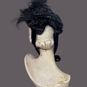 May include: A vintage black hat with sequins and feathers, displayed on a white bust. The hat features a decorative bow and long black ribbons. The hat's design is intricate, with a focus on texture and detail.