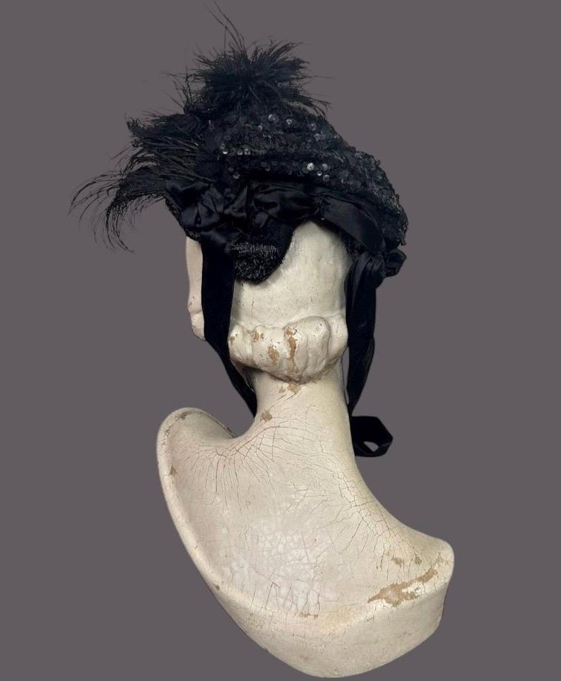 May include: A vintage black hat with sequins and feathers, displayed on a white bust. The hat features a decorative bow and long black ribbons. The hat's design is intricate, with a focus on texture and detail.
