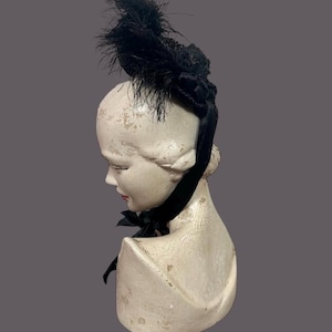 May include: A black feather hat with a decorative bow and ribbon, displayed on a vintage mannequin head. The hat features a cluster of black feathers and a velvet ribbon that ties into a bow. The mannequin head is off-white with a distressed finish.