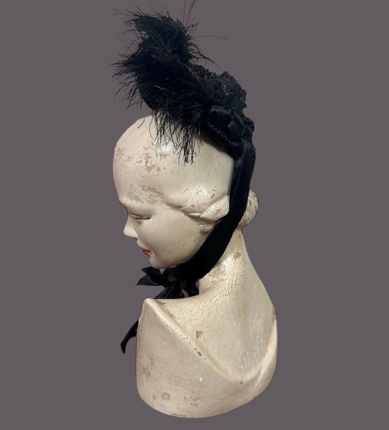 May include: A black feather hat with a decorative bow and ribbon, displayed on a vintage mannequin head. The hat features a cluster of black feathers and a velvet ribbon that ties into a bow. The mannequin head is off-white with a distressed finish.