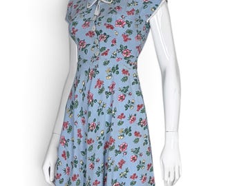 May include: A light blue knee-length dress featuring a floral pattern with pink, red, and yellow flowers and green leaves. The dress has short sleeves, a high collar with a white trim, and a front button closure with a decorative tie at the neck.